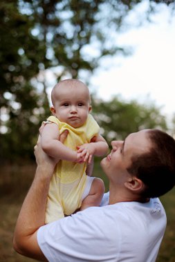 Baba ve küçük bebek doğayla oynayıp gülüyorlar. Babalar Günü. Küçük kızını ağaçların arasında, doğada oynarken kollarında tutuyor. Baba sarılıp öper sarışın çocuğunu.