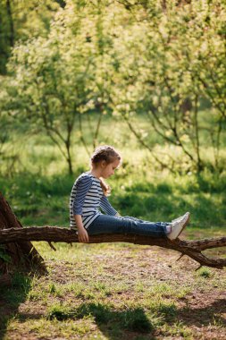 Güneşli bir günde devrilmiş bir ağaçta oturan küçük bir kız yeşil bir ormanın arka planında. Çizgili elbiseli ve kot pantolonlu kız. Çocukluk eğlenceleri ve oyunlar, açık hava aktiviteleri konsepti. Seçici odak.