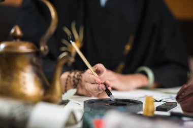 Siyah gömlekli bir adam geleneksel Çin kaligrafisi için fırçasını mürekkebe batırıyor. Ön planda çözünürlükte bakır bir su ısıtıcısı var. Masanın üzerinde hiyeroglifli kağıt var. yakın plan.