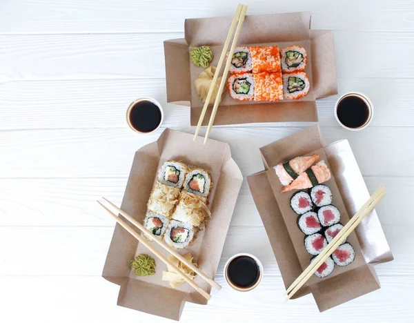 Flat lay composition with sushi rolls, chopsticks, marinated ginger, soy sauce, wasabi in eco boxes on white wooden table. Food delivery and takeaway concept