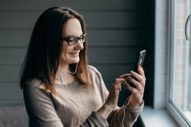Gözlüklü mutlu esmer kadın evde akıllı telefonuyla görüntülü konuşma yapıyor, yakınlaştırma toplantısı uygulaması kullanıyor, sosyal uzaklık, evden çalışma, uzaktan konsept çalışıyor.