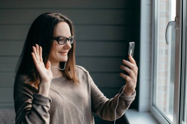 Gözlüklü mutlu esmer kadın evde akıllı telefonuyla görüntülü konuşma yapıyor, yakınlaştırma toplantısı uygulaması kullanıyor, sosyal uzaklık, evden çalışma, uzaktan konsept çalışıyor.