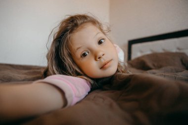 Covid-19 karantinası sırasında çocuk pijama partisinde iyi eğlenceler. Yatakta yatıp kameraya bakan komik küçük kız.