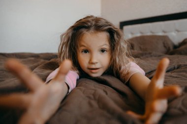 Covid-19 karantinası sırasında çocuk pijama partisinde iyi eğlenceler. Yatakta uzanmış, omuz silkip sürpriz yapan komik küçük kız.