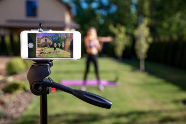 Sportive fitness coach woman recording video blog  by phone about  workout  training at the garden with fresh green grass near house.  clipart