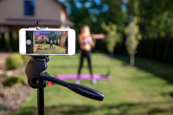 Sportive fitness coach woman recording video blog  by phone about  workout  training at the garden with fresh green grass near house. 