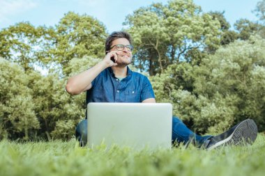 Çimenlerin üzerinde otururken Smartphone kullanan genç adam