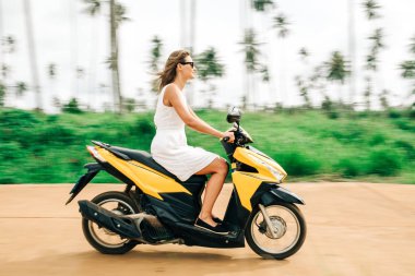 Beyaz elbiseli, güneş gözlüklü, motosiklet süren mutlu kadın.
