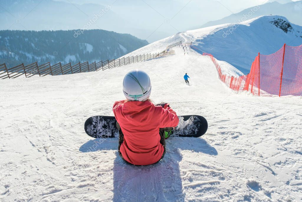 Snowboarder girl sits with board at the ski slope in mountains