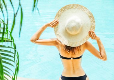 Güzel bir kadın şapkaya dokunuyor ve tatil köyünde havuzun keyfini çıkarıyor.