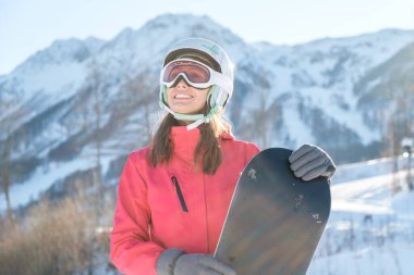 Kasklı ve maskeli bir sporcunun portresi elinde kar kaykayıyla kameraya bakıyor, gülümsüyor, güneşli dondurucu günün tadını çıkarıyor.