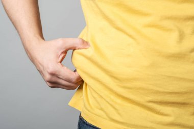 Şişman, şişman bir adamın, koca göbeğini tutarak gri arka plan üzerinde izole bir şekilde elini tutmasına yakın durun.