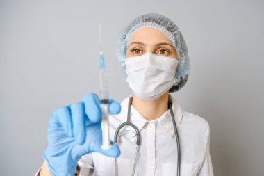 Tıbbi üniformalı, koruyucu maskeli ve elinde şırınga olan genç bir kadının portresi. Gri arkaplanda izole