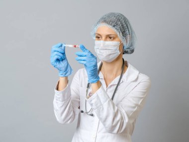 Koruyucu maskeli ve eldivenli genç çekici kadın bilim adamı test tüpündeki kırmızı sıvı maddeye bakıyor. Gri arkaplanda izole
