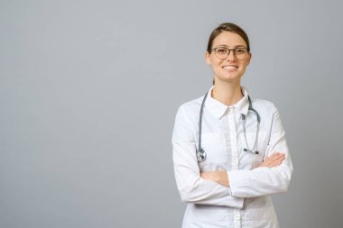 Kolları gri arka planda çaprazlanmış laboratuvar önlüklü gülümseyen kadın doktor.