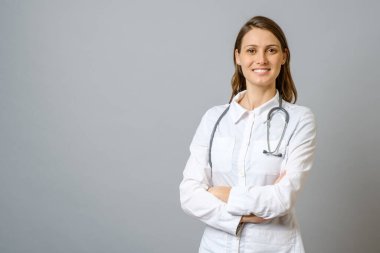 Kolları gri arka planda çaprazlanmış laboratuvar önlüklü gülümseyen kadın doktor.