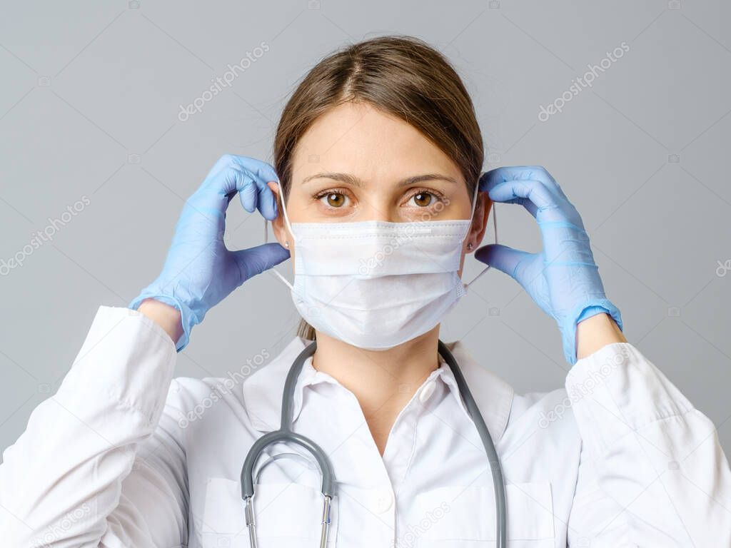 Portrait of a young female medical doctor or nurse putting sterile mask isolated on gray background