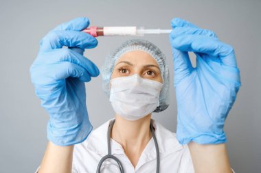 Koruyucu maskeli ve eldivenli genç çekici kadın bilim adamı test tüpündeki kırmızı sıvı maddeye bakıyor. Gri arkaplanda izole