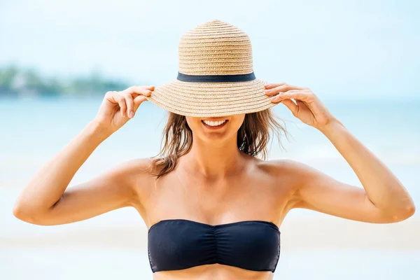 Beautiful tanned woman in black bikini covering face with a straw hat, posing on the tropical beach
