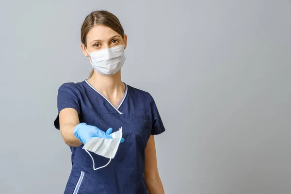 青いコートとフェイスマスクを身に着けている若い女性医師は フェイスマスクを提供しています 灰色の背景に隔離された — ストック写真
