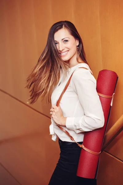 Portrait of beautiful smiling young white Caucasian business woman with blue eyes in white blouse shirt and black pants holding, carrying yoga mat in office, standing on staircase, lifestyle concept — Stock Photo, Image