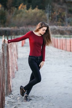 dalgın hüzünlü yalnız beyaz genç güzel bir kadın dağınık uzun saçlı rüzgarlı gün açık gün batımında ahşap çit yakınındaki kıyı sahilde portresi