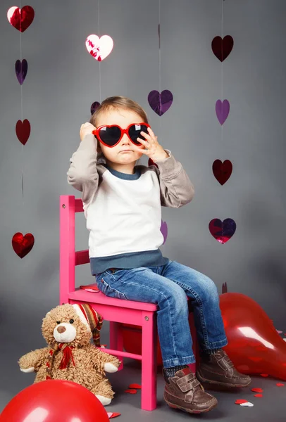 Porträtt av vita kaukasiska söt bedårande lilla baby pojke småbarn sitter på små rosa stol med Björn leksak i studio bär röda roliga glasögon leende ha roligt, semester alla hjärtans dag — Stockfoto