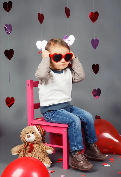 Portrait of white Caucasian cute adorable little baby boy toddler sitting on small pink chair with bear toy in studio wearing red funny glasses smile having fun, holiday Valentine day — Stok Foto