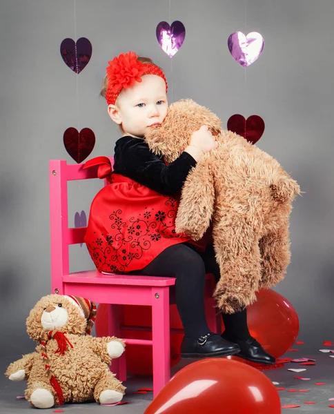 Portrait of white Caucasian cute adorable little baby girl toddler sitting on small pink chair with bear toy in studio wearing red dress and headband, holiday Valentine day