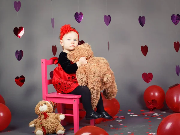 Retrato de blanco caucásico lindo adorable bebé niña niño sentado en una pequeña silla rosa con juguete oso en el estudio con vestido rojo y diadema, día de San Valentín de vacaciones — Foto de Stock