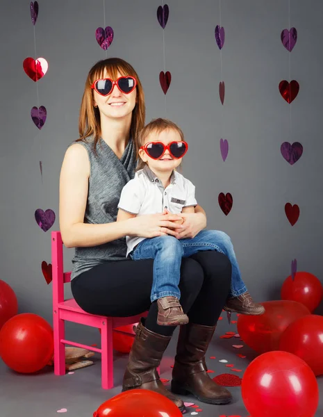 Портрет молодой кавказской матери, сидящей на маленьком розовом стульчике с маленьким мальчиком малышом на коленях в студии в смешных очках, улыбающейся смеясь, веселясь, праздник День Святого Валентина — стоковое фото
