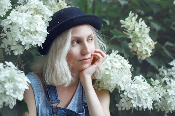 Closeup portret van mooie Kaukasische teenage jonge blonde alternatief model meisje vrouw in blauw t-shirt, jeans romper, zwarte hoed, zittend onder grote witte bloemen op een zomerdag — Stockfoto