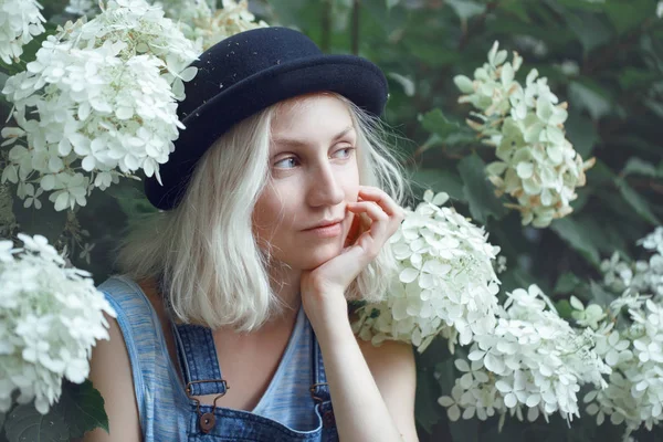 Gros plan portrait de belle adolescente caucasienne jeune fille modèle alternatif blonde femme en tshirt bleu, jean barboteuse, chapeau noir, assis parmi de grandes fleurs blanches le jour de l'été — Photo