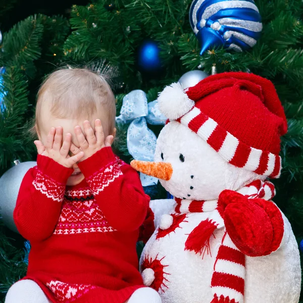 Portrait de petite petite fille blanche tout-petit en robe rouge assis près de l'arbre du Nouvel An avec jouet bonhomme de neige fermeture couvrant ses yeux avec les mains dans la surprise, le bonheur de l'enfance — Photo