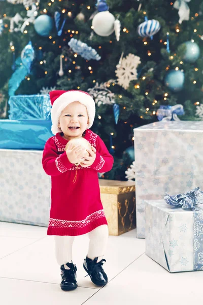 Portrait de bébé fille caucasienne souriante heureuse tout-petit avec les yeux bleus dans la robe rouge et le chapeau du Père Noël debout près de l'arbre du Nouvel An près des boîtes-cadeaux tenant décoration d'ornement d'or, image de Noël style de vie . — Photo