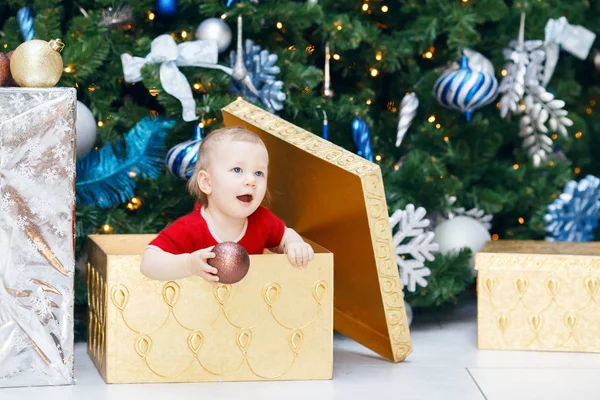 Portrait of happy funny smiling Caucasian baby girl toddler in red holiday dress sitting in large gift present box  under New Year tree holding ornaments decoration. — Stock Photo, Image