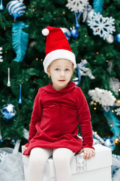 Portrait de sourire heureux enfant caucasien drôle fille tout-petit avec les yeux bleus en robe rouge et le chapeau du Père Noël assis sur une boîte par arbre de Nouvel An, concept de vacances de Noël style de vie — Photo