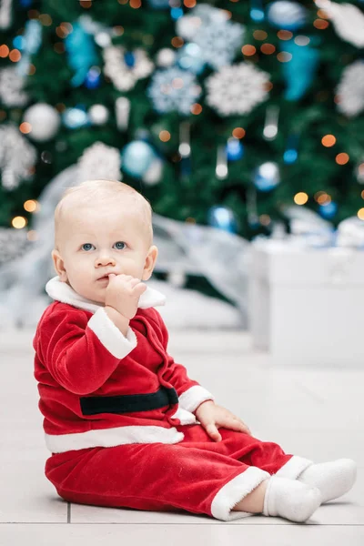 Portrait de bébé garçon caucasien blond aux yeux bleus en costume rouge du Père Noël assis près de l'arbre du Nouvel An, concept de vacances de Noël style de vie — Photo