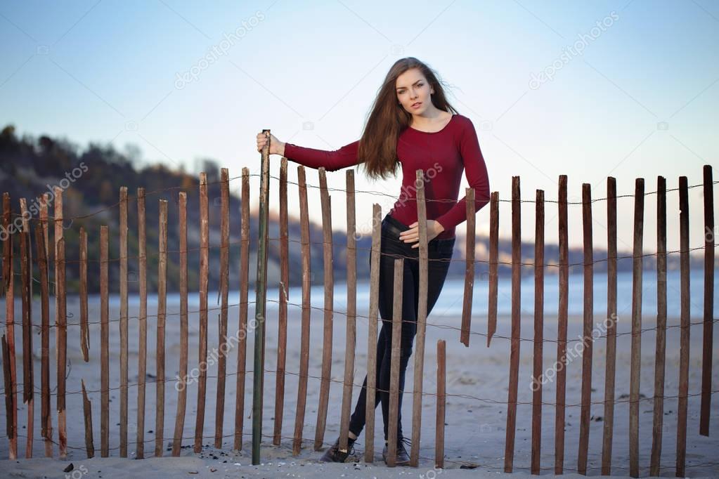 portrait of pensive sad lonely Caucasian young beautiful woman with messy long hair on windy day outdoor on the shore beach near wooden fence at sunset