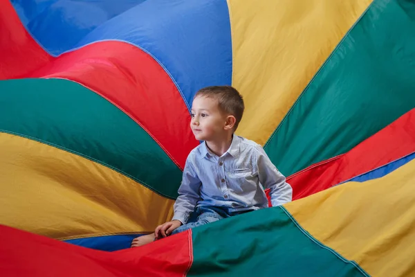 Retrato de niño blanco caucásico niño sentado en el centro del parque infantil paracaídas de arco iris celebrando su cumpleaños en la fiesta — Foto de Stock