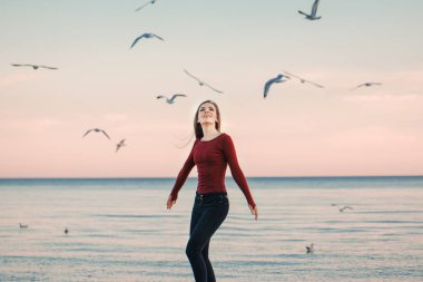 Beyaz martılar kuşlar arasında sonbahar sonbahar gününde açık kıyı sahilde günbatımı, yaşam tarzı doğal duygu atlama koşma kot pantolon bir genç kadın mutlu gülümseyen gülen portre heyecanlı