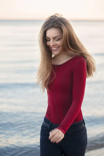 Retrato de estilo de vida de la joven mujer delgada caucásica con el pelo largo desordenado usando jeans negros y camisa roja caminando en el día ventoso al aire libre en la orilla de la playa al atardecer, desenfoque de acción de movimiento —  Fotos de Stock