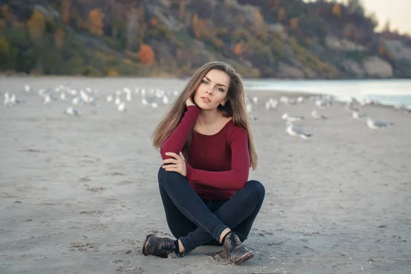 Portret van de levensstijl van de Kaukasische jonge mooie vrouw met lang haar, in zwarte jeans en rood shirt, vergadering op zand op strand onder meeuwen vogels op herfst vallen dag buiten bij zonsondergang — Stockfoto