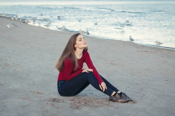 Lifestyle portrait of Caucasian young beautiful woman with closed eyes, long hair, in black jeans and red shirt, sitting on sand on beach among seagulls birds on autumn fall sunset — Stock Photo, Image