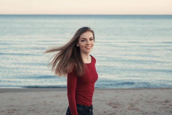 Retrato de feliz sonrisa riendo despreocupado blanco caucásico joven hermosa mujer con pelo largo desordenado en el día ventoso al aire libre al atardecer en la playa por el agua, mirando en la cámara — Foto de Stock