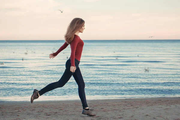 Portrait of happy smiling laughing excited Caucasian young woman in jeans running jumping among seagulls birds on autumn fall day outdoor on the shore beach at sunset, lifestyle natural emotion — Stock Photo, Image