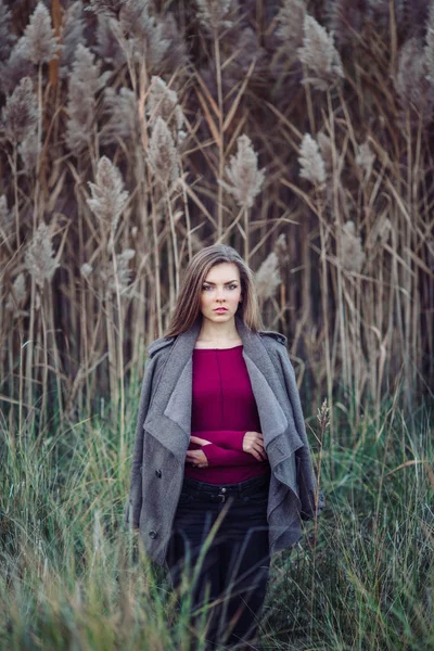 Portrait of pensive sad lonely Caucasian blonde young beautiful woman girl with long hair wearing jeans, coat jacket,  in forest field among large tall plants grass looking in camera — Stock Photo, Image