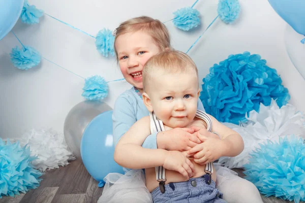 Portrait de deux souriants embrassant mignon adorable enfants caucasiens, petite fille et bébé garçon, célébrant anniversaire regardant à la caméra, fond studio blanc avec des ballons bleus — Photo