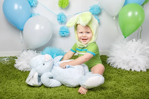 Portrait of cute adorable Caucasian baby boy in holiday Easter bunny rabbit costume with large ears,  dressed in green clothes onesie, sitting on soft fluffy rug carpet in studio, holding red carrot — Stock Photo, Image