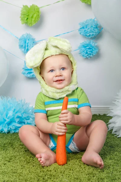 Portrait de mignon adorable bébé garçon caucasien en vacances Lapin de Pâques costume de lapin avec de grandes oreilles, vêtu de vêtements verts onesie, assis sur tapis moelleux moelleux en studio, tenant carotte rouge — Photo